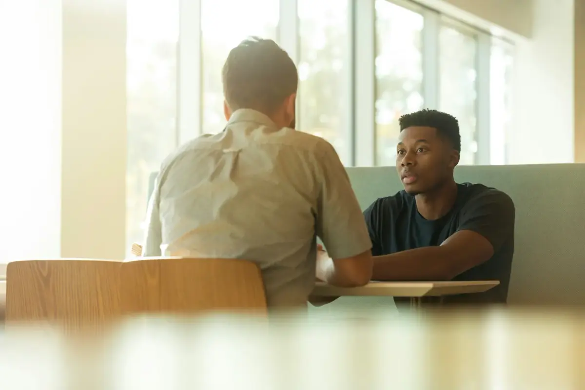 Two men in a serious one-on-one conversation at a small table by a sunny window
