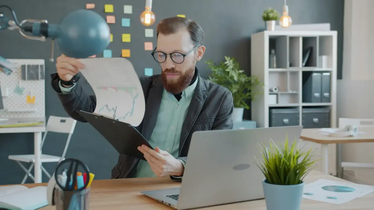 A man at his office desk holding up a printed document to read it, a laptop open in front of him