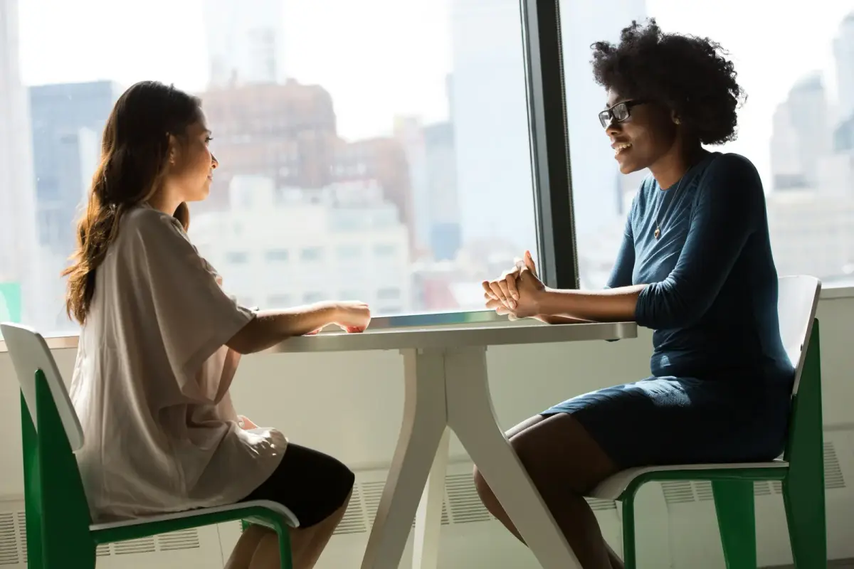 Two women sitting across a small table by an office window, talking one-on-one
