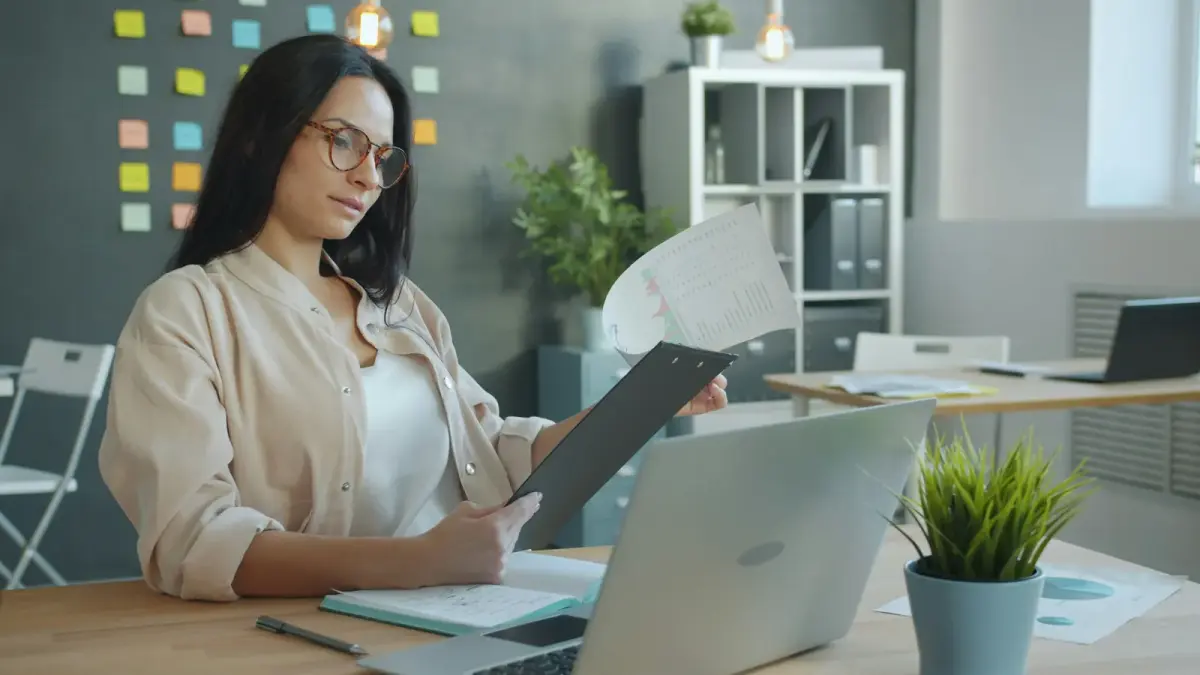 A woman at an office desk reading a document on a clipboard beside her laptop
