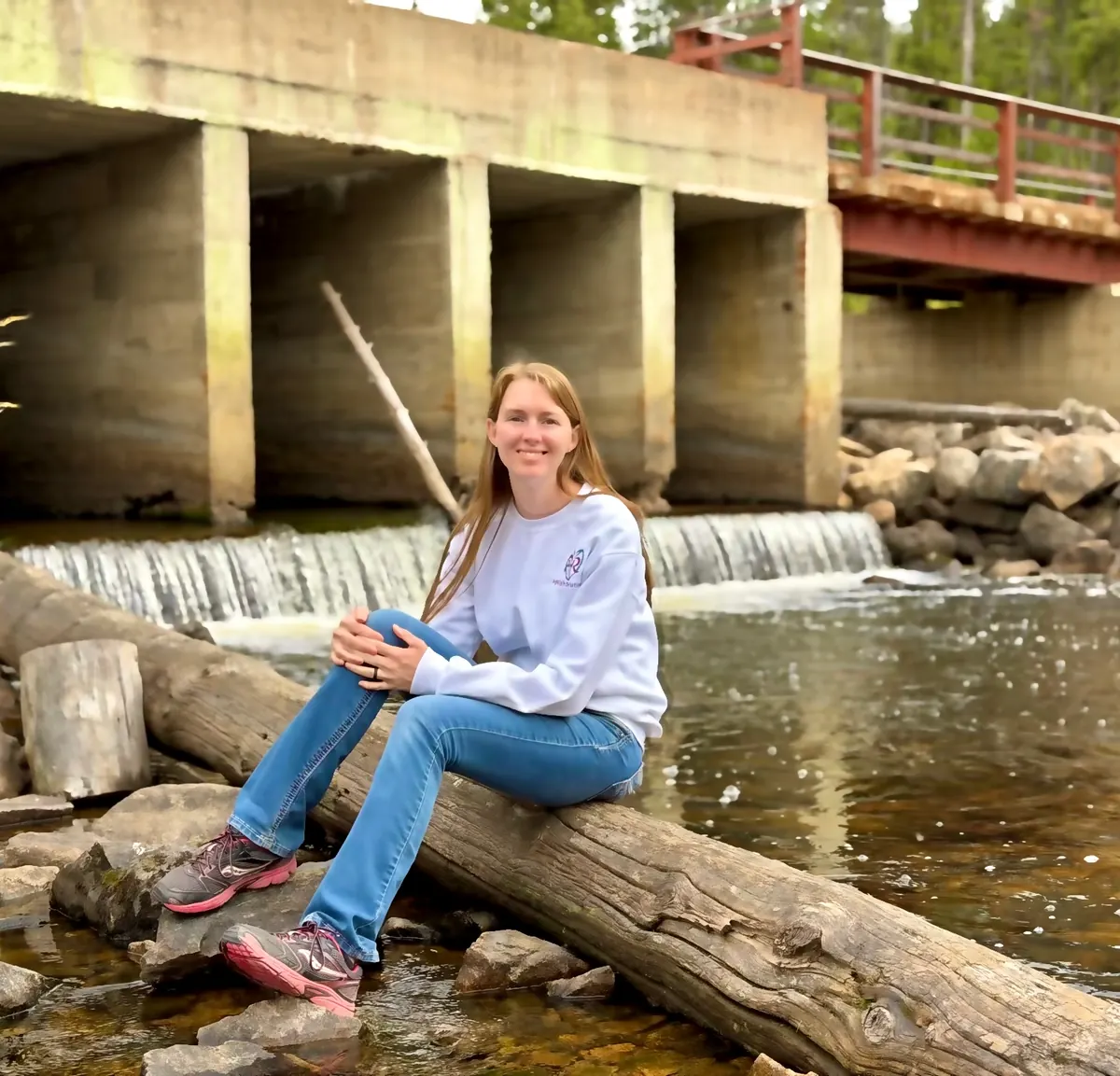 Allisha McCartney sitting on a log by a river, below a small waterfall and a bridge