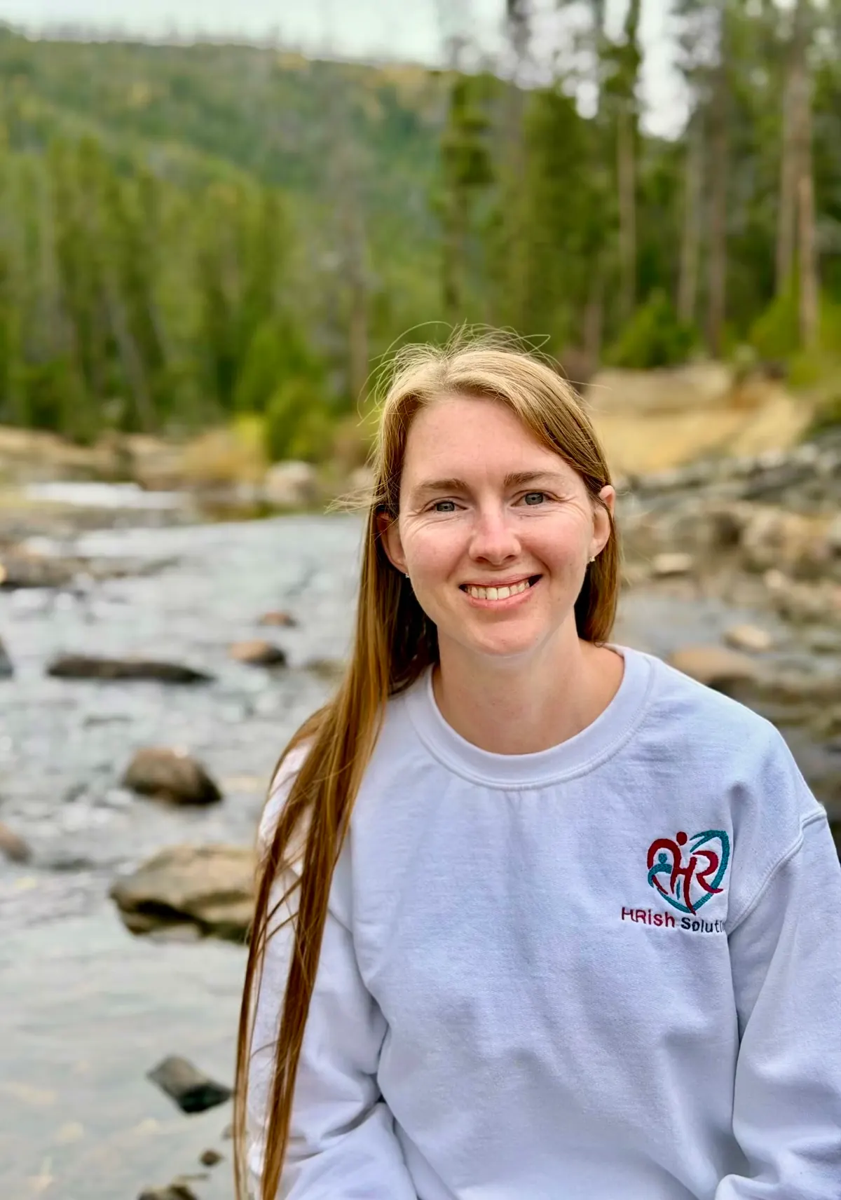 Allisha McCartney smiling by a mountain river in an HRish Solutions sweatshirt