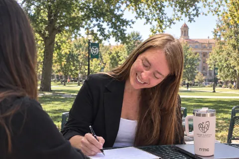 Allisha McCartney smiling and writing in a notebook at an outdoor table on the Colorado State University campus
