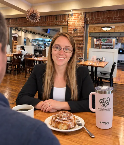 Allisha McCartney smiling across a café table, with a coffee, a cinnamon roll and her HRish Solutions tumbler